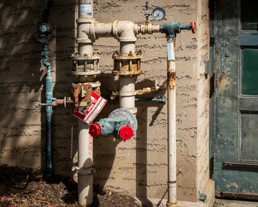 Rusty outdoor plumbing pipes with pressure gauges and warning signs on a wall.